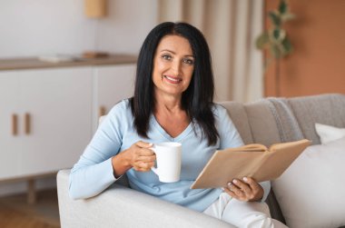 Kitap okuyan ve elinde kahve tutan olgun bir kadın.