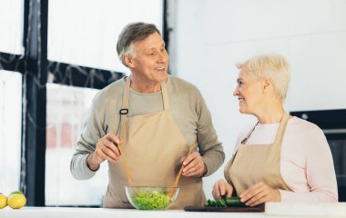 Kıdemli çift mutfakta yemek pişiriyor, gülümsüyor.