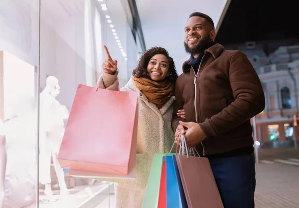 Feliz pareja negra con bolsas de compras apuntando a la ventana del centro comercial —  Fotos de Stock