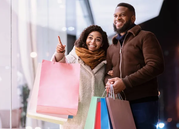 Feliz pareja negra con bolsas de compras apuntando a la ventana del centro comercial —  Fotos de Stock
