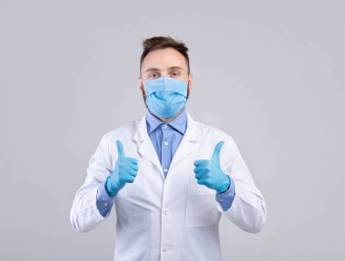 Üniformalı, maskeli ve eldivenli milenyum doktoru kameraya bakıyor ve gri arkaplan üzerinde baş parmak işareti yapıyor.