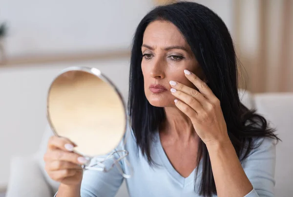 Femme mûre vérifiant son visage dans le miroir, toucher le visage — Photo