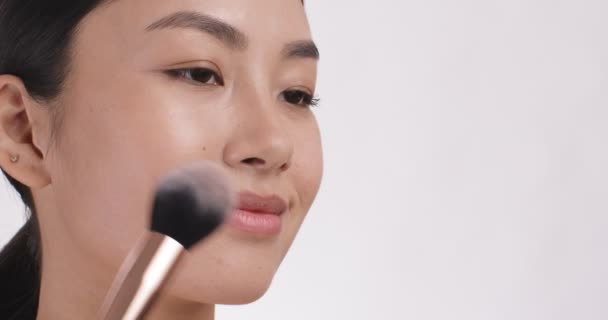 Joven mujer asiática aplicando rubor en la cara con cepillo de maquillaje. Concepto cosmético, primer plano, fondo blanco del estudio — Vídeo de stock