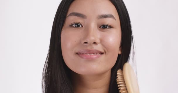 Joven feliz asiática señora peinándose el pelo con cepillo derecho, sonriendo a la cámara, fondo blanco estudio, primer plano retrato — Vídeo de stock