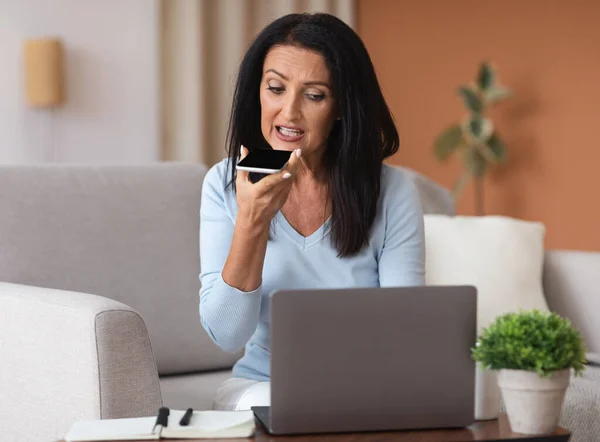 Mulher madura usando laptop e assistente de voz em seu smartphone — Fotografia de Stock