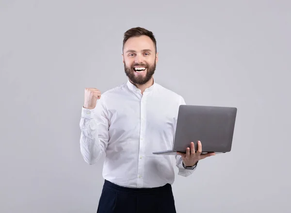 Захоплений офісний працівник у формальному носінні тримає ноутбук і робить жест YES над сірим студійним фоном — стокове фото