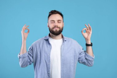 Stres yönetimi. Huzurlu genç adam gözleri kapalı meditasyon yapıyor, elleri mavi arka planda Gyan Mudra yapıyor.