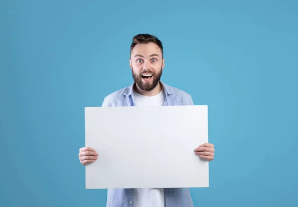 Aufgeregter junger Mann mit leerem Poster mit Kopierraum auf blauem Studiohintergrund — Stockfoto