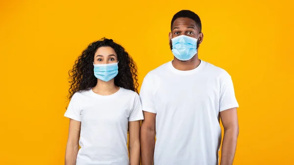 Casal multiétnico vestindo máscaras faciais posando sobre fundo de estúdio amarelo — Fotografia de Stock