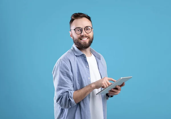 Позитивний молодий хлопець у повсякденному носінні використовує планшетний комп'ютер на синьому студійному фоні — стокове фото
