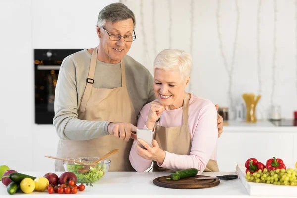 Alegre cónyuges mayores usando teléfono celular cocina cena en la cocina —  Fotos de Stock