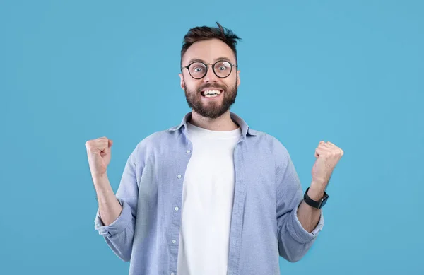 Захоплений бородатий хлопець робить YES жест, посміхаючись, стискаючи кулаки на синьому студійному фоні, панорама — стокове фото