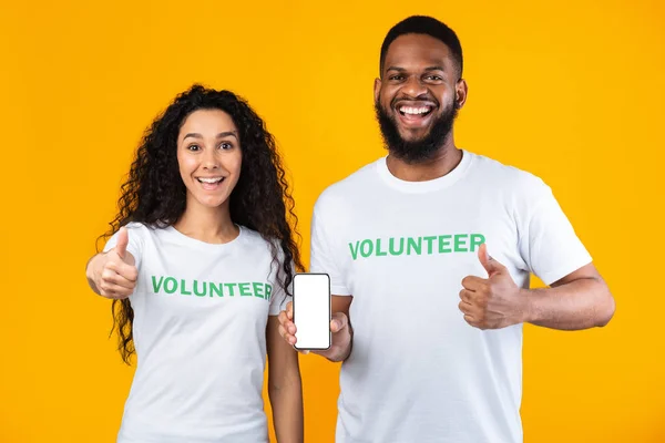 Two Volunteers Showing Smartphone Screen Gesturing Thumbs-Up On Yellow Background — Stock Photo, Image