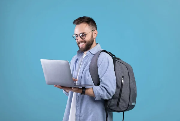 Cool millennial cara com mochila usando laptop para trabalho on-line ou estudos sobre fundo estúdio azul — Fotografia de Stock