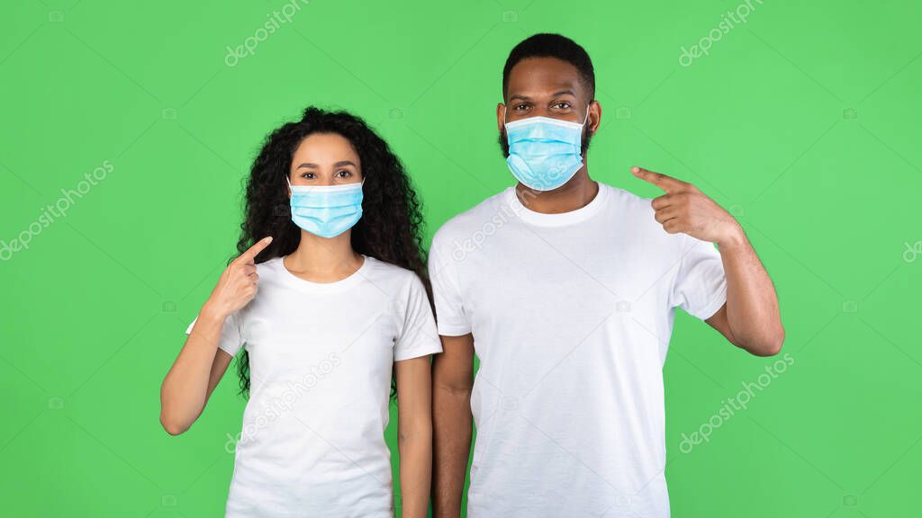Multiracial Couple Pointing Fingers At Masks On Face, Green Background
