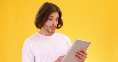 Dijital tablet hakkında bilgi okuyan genç bir adamın stüdyo portresi, cihaz, sarı arka plan, yavaş çekim