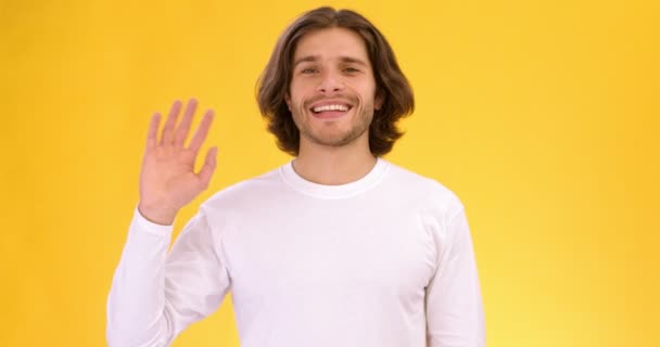 Studio shot van jonge vriendelijke spraakzame man zwaaiende hand en zeggen Hallo, slow motion, oranje achtergrond — Stockvideo