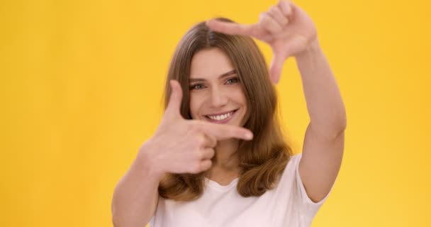 Studio portret van vrolijke jonge dame inlijsten haar gezicht met vingers en glimlachen om de camera, oranje achtergrond — Stockvideo