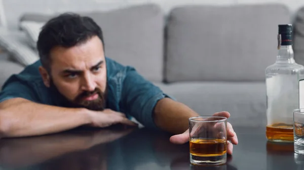 Gefrustreerde man met baard neemt een glas whisky van tafel in de woonkamer interieur — Stockfoto