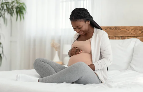 Щаслива чорна очікувана леді насолоджується вагітністю, сидячи на ліжку торкаючись живота — стокове фото