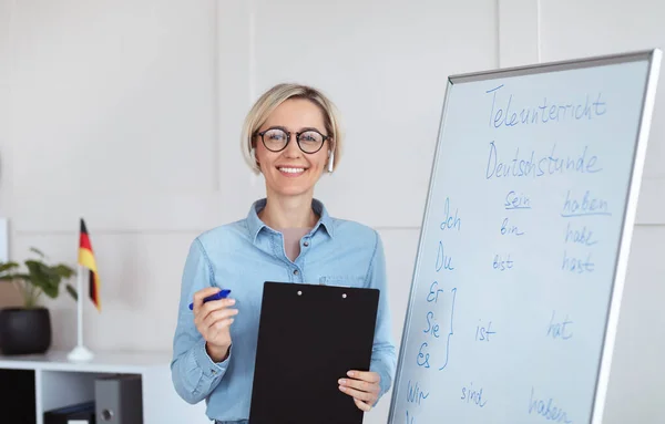Çevrimiçi eğitim. Gramer kurallarıyla tahtaya yakın poz veren pozitif Almanca öğretmeni kameraya bakıyor ve gülümsüyor. — Stok fotoğraf