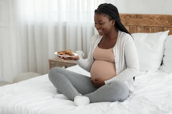 Černá těhotná žena s talířem sušenek sedí doma na posteli — Stock fotografie