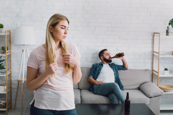 Сумна дружина бачить, як її чоловік п'є багато пива, чоловік з бородою сидить на дивані — стокове фото