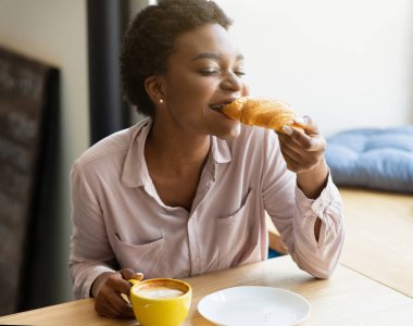 Güzel, genç, siyah bir kadın şehir kafesinde kruvasan ve kahvenin tadını çıkarıyor.