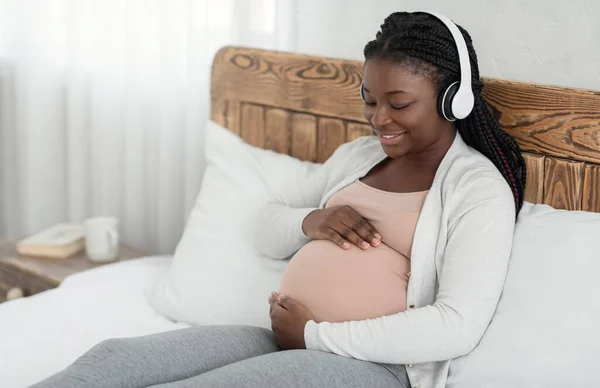 Güzel siyah hamile kadın, kablosuz kulaklıkla müzik dinliyor ve karnına dokunuyor. — Stok fotoğraf