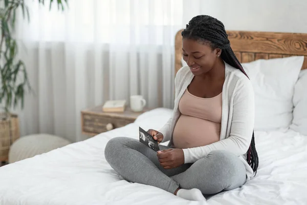 Feliz mujer embarazada africana sentada en la cama con imagen de exploración por ultrasonido de bebé —  Fotos de Stock