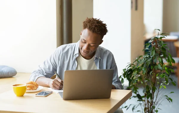 Slimme zwarte man met laptop werken aan project of studeren op afstand, schrijven in notebook in gezellige stedelijke cafe — Stockfoto