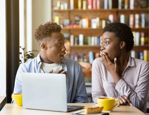 Jovem mulher negra cobrindo a boca em choque e seu namorado surpreso perto de computador portátil no café urbano — Fotografia de Stock