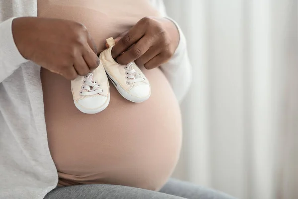 Nerozpoznatelná černá těhotná dáma drží malé dětské boty v blízkosti svého břicha — Stock fotografie