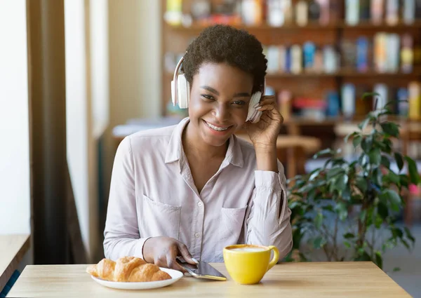 Retrato de una encantadora dama negra con teléfono móvil con auriculares en la cafetería urbana, disfrutando de música o audiolibro —  Fotos de Stock