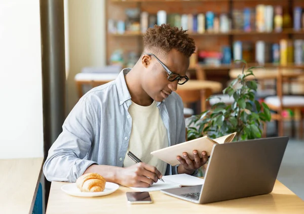 Bonito negro de óculos aprendendo remotamente no laptop, tomando notas no caderno, estudando on-line no café da cidade — Fotografia de Stock