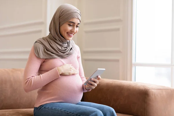 Šťastná těhotná muslimská žena pomocí chytrého telefonu sedí na pohovce vnitřní — Stock fotografie