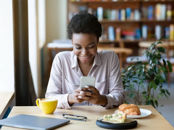 Joven mujer afroamericana con teléfono inteligente chatear en línea, navegar por la web, comprobar las redes sociales en la cafetería — Foto de Stock