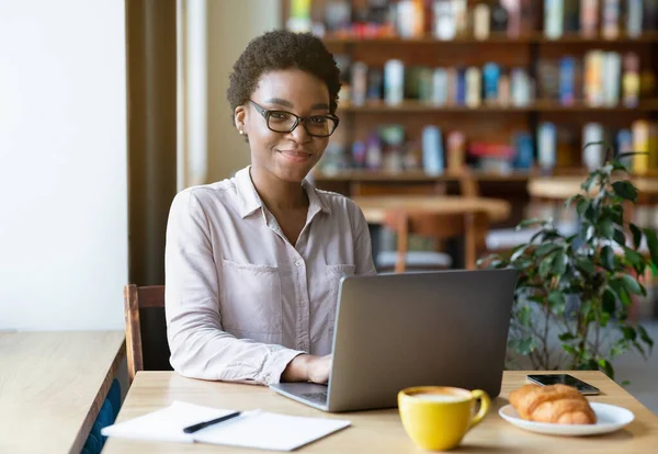 Távoktatás és online munka koncepció. Okos fiatal hölgy szemüveg segítségével laptop számítógép hangulatos városi kávézó — Stock Fotó