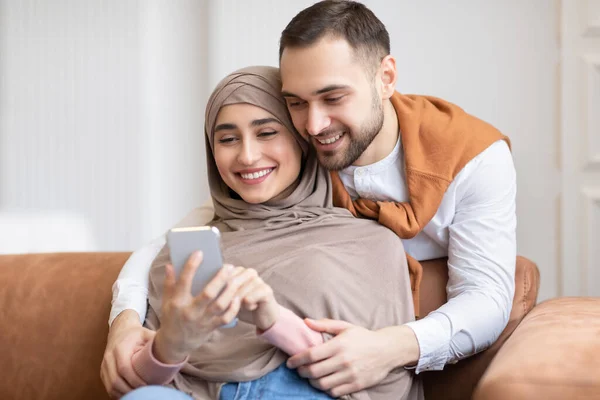 Casal muçulmano feliz usando mensagens de texto Smartphone sentado no sofá dentro de casa — Fotografia de Stock