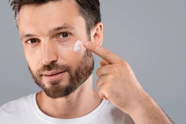 Fechar-se de homem barbudo aplicando creme facial no cinza — Fotografia de Stock