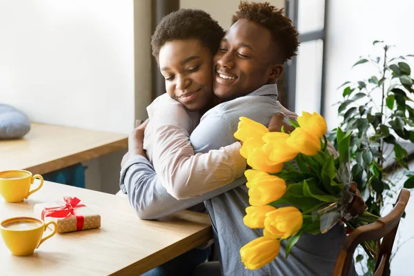 Couple noir affectueux avec des fleurs jaunes et des cadeaux s'embrassant au café, célébrant ensemble la Saint-Valentin — Photo