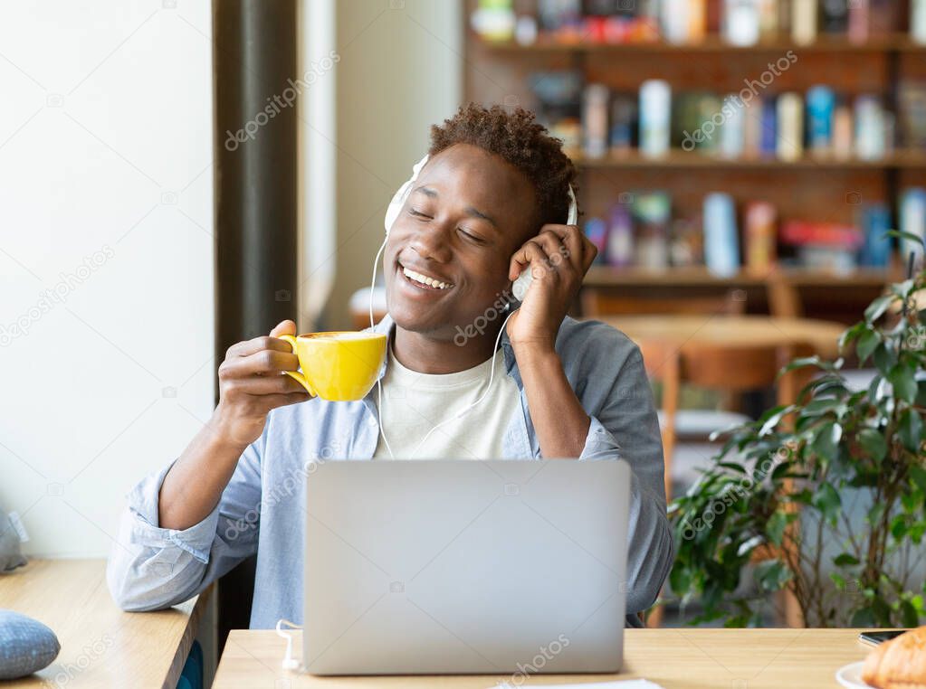 Handsome black guy with headphones listening to music with closed eyes and drinking coffee at cozy cafe