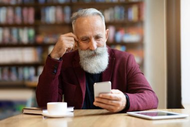 Yaşlı gri saçlı adam kafede kahve içiyor, akıllı telefon kullanıyor.