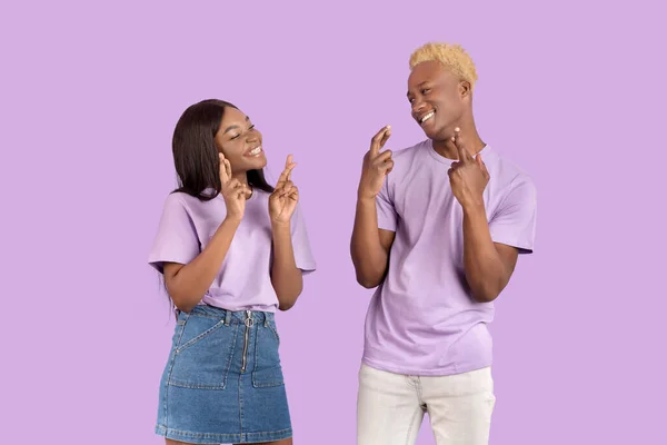 Chico negro alegre y su novia pidiendo deseo con los dedos cruzados, rogando buena suerte sobre fondo violeta — Foto de Stock