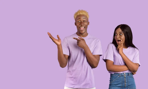 Mujer afroamericana y su novio se sienten conmocionados, señalando el espacio en blanco sobre fondo lila, diseño de pancartas — Foto de Stock