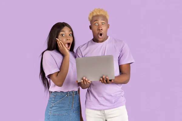 Millennial casal preto com laptop olhando para a câmera em choque, de pé com bocas abertas sobre violeta estúdio fundo — Fotografia de Stock