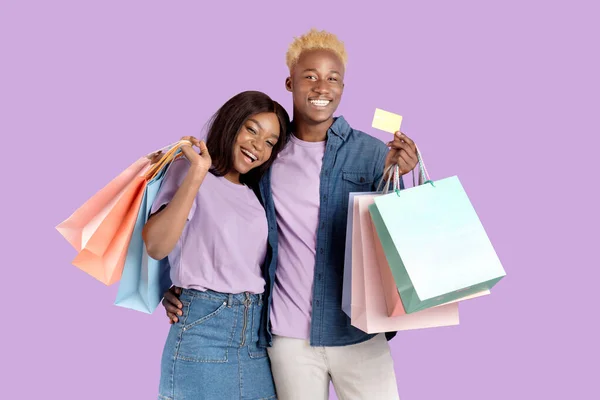 Afectuosa pareja negra sosteniendo bolsas de regalo y tarjeta de crédito, promoviendo el pago sin contacto sobre fondo violeta —  Fotos de Stock