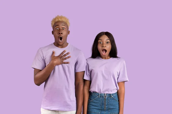 Impresionante pareja afroamericana abriendo bocas en shock, poniendo la mano en el corazón sobre fondo violeta estudio — Foto de Stock