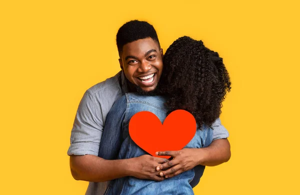 Romantico uomo di colore che abbraccia la fidanzata e tiene il cuore di carta rossa, facendo sorpresa — Foto Stock