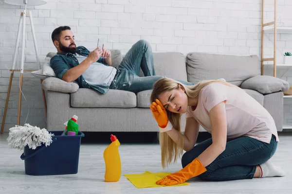 Frau leidet unter Kopfschmerzen und putzt zu Hause, Mann mit Bart liegt auf Couch und spielt am Telefon — Stockfoto